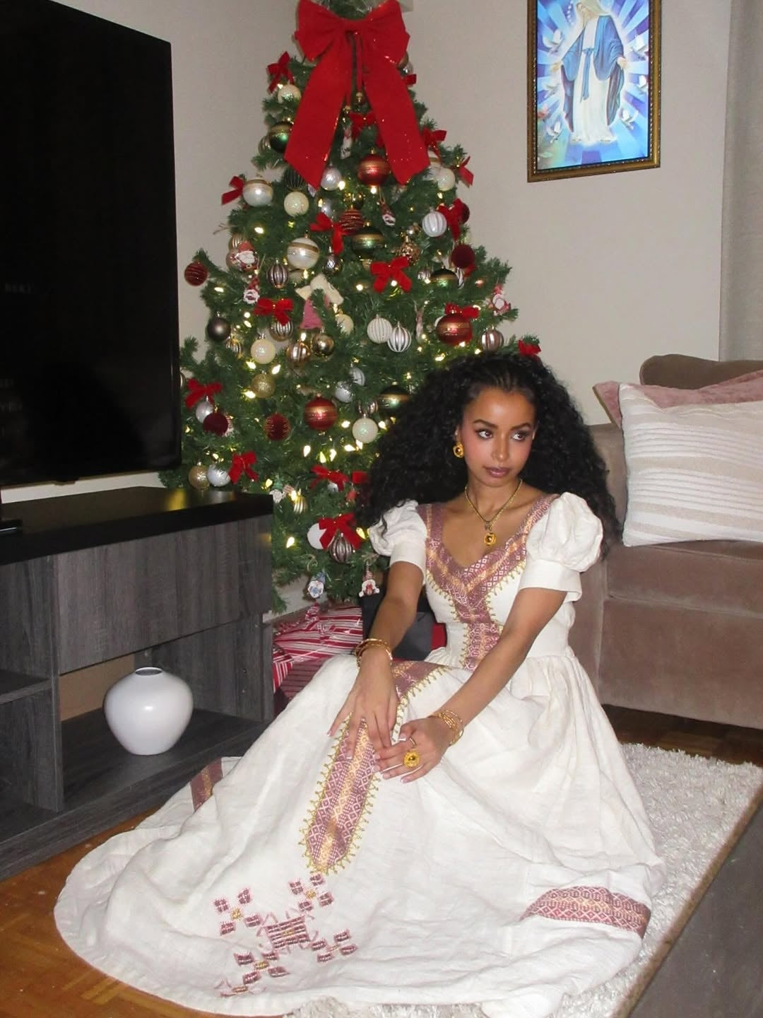 Woman in a white Habesha Libs sitting in front of a decorated Christmas tree in a living room.