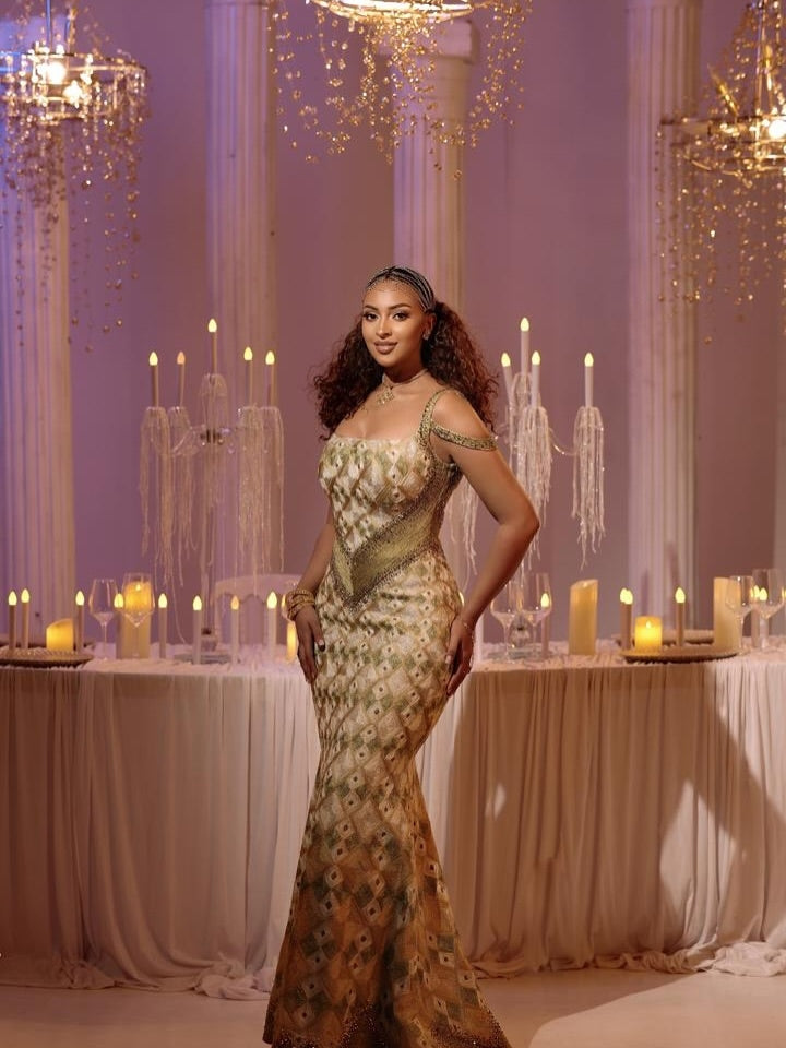 A woman wearing a gold and beige embellished  Ethiopian Dress  sits on a draped sofa in a warmly lit event setting with tables, candles, and hanging lights in the background.