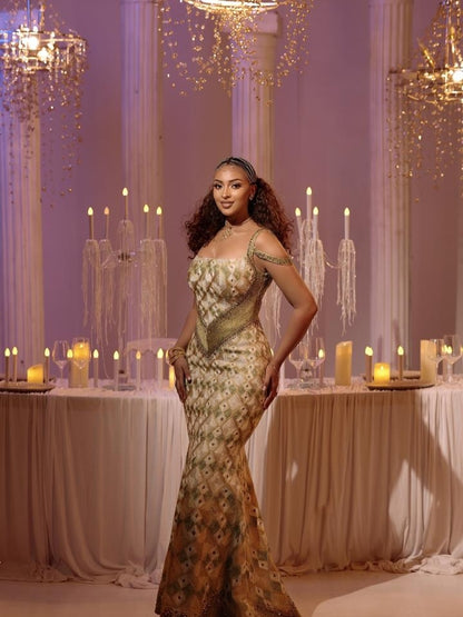 A woman wearing a gold and beige embellished  Ethiopian Dress  sits on a draped sofa in a warmly lit event setting with tables, candles, and hanging lights in the background.
