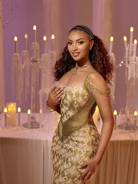 A woman wearing a gold and beige embellished  Ethiopian Dress  sits on a draped sofa in a warmly lit event setting with tables, candles, and hanging lights in the background.