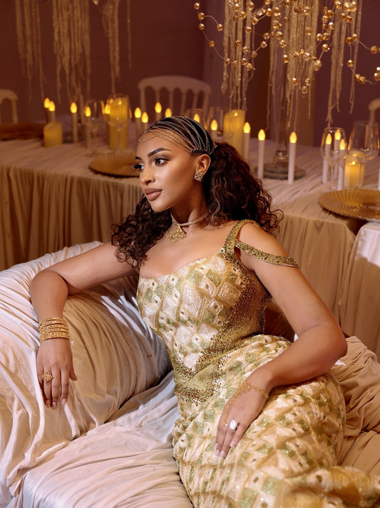 A woman wearing a gold and beige embellished  Ethiopian Dress  sits on a draped sofa in a warmly lit event setting with tables, candles, and hanging lights in the background.