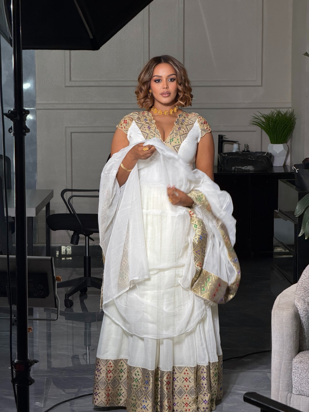 Woman in a white and gold traditional Habesha Kemis holding a white shawl in a room.