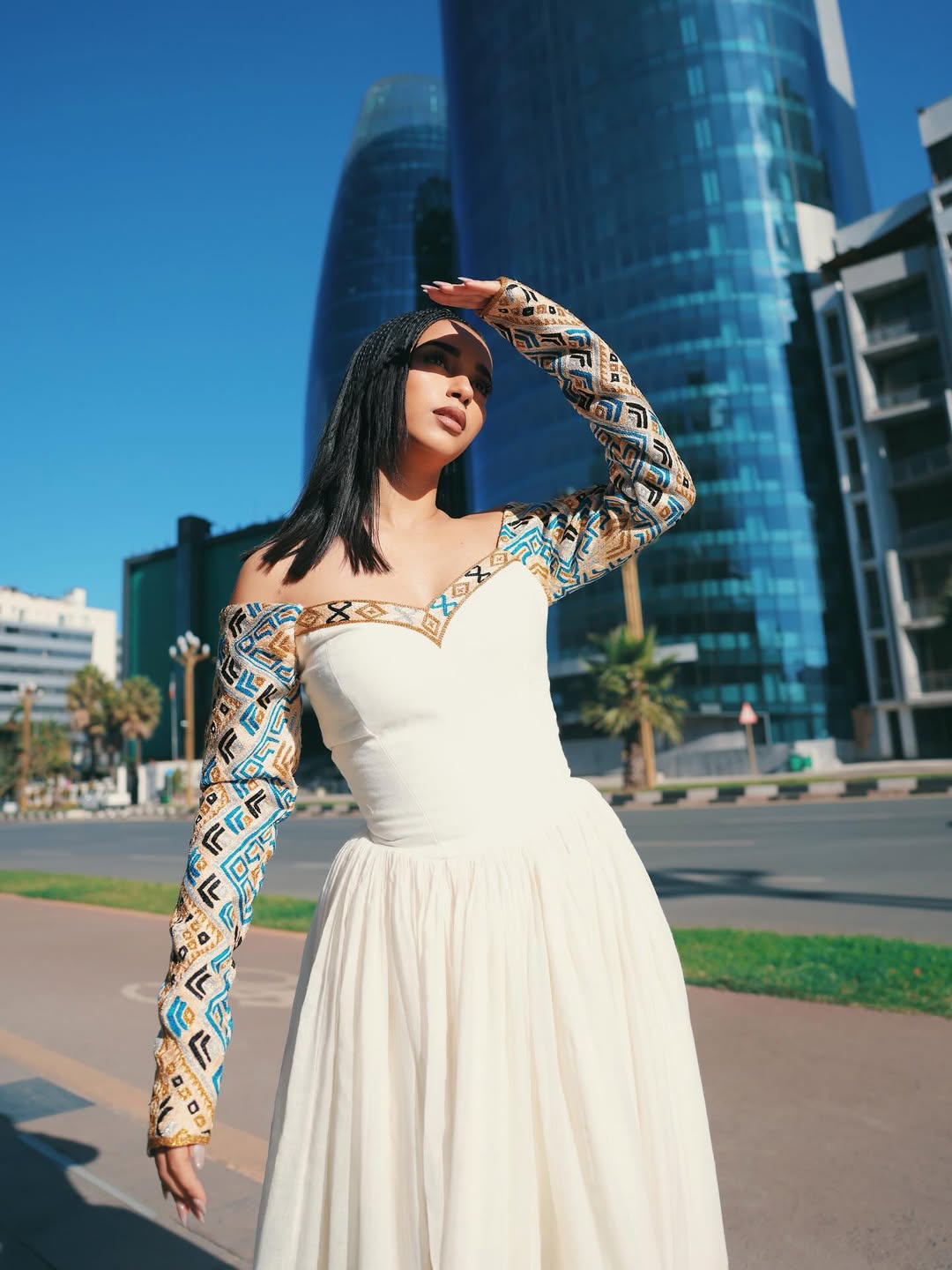 Woman in a white Ethiopian dress with patterned sleeves standing in front of modern buildings.