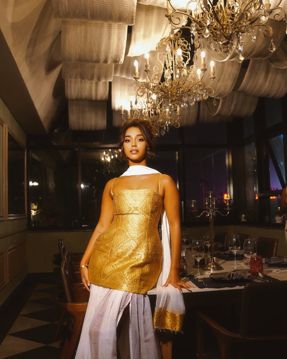 A woman stands in a dimly lit restaurant or event space wearing a metallic gold sleeveless Habesha Dress  with a white draped scarf and light lavender skirt. Large chandeliers, tables, and dark windows are visible in the background.