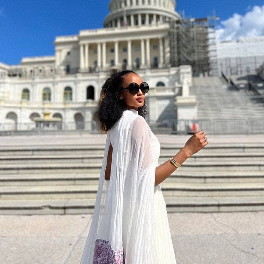 A woman wearing a white draped Ethiopian Dress  and black sunglasses stands in front of the U.S. Capitol building on a sunny day.