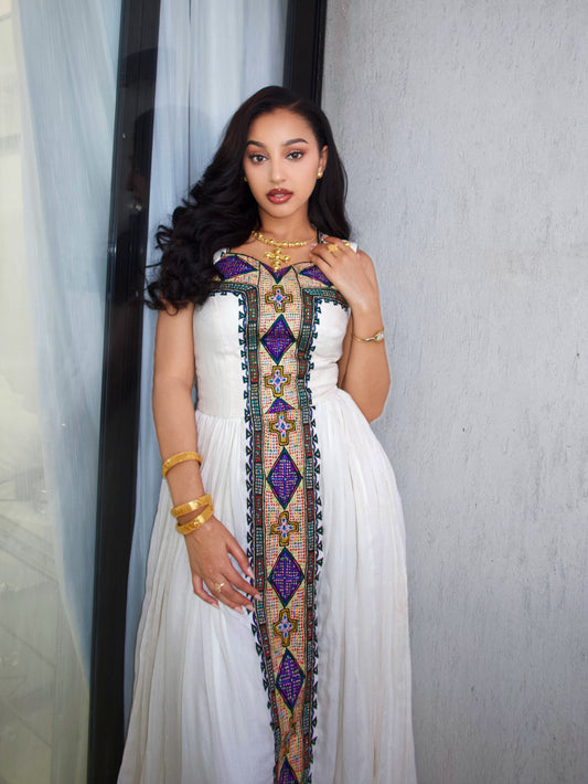 A woman wearing a white sleeveless Ethiopia Clothes with a prominent vertical purple, gold, and green geometric embroidered panel down the front, accessorized with gold jewelry, standing against a textured gray wall.