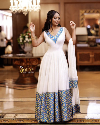 A woman modeling a white floor-length Habesha Kemis  with blue and gold geometric patterns on the neckline and hem, paired with a matching white dupatta, in an indoor event space.