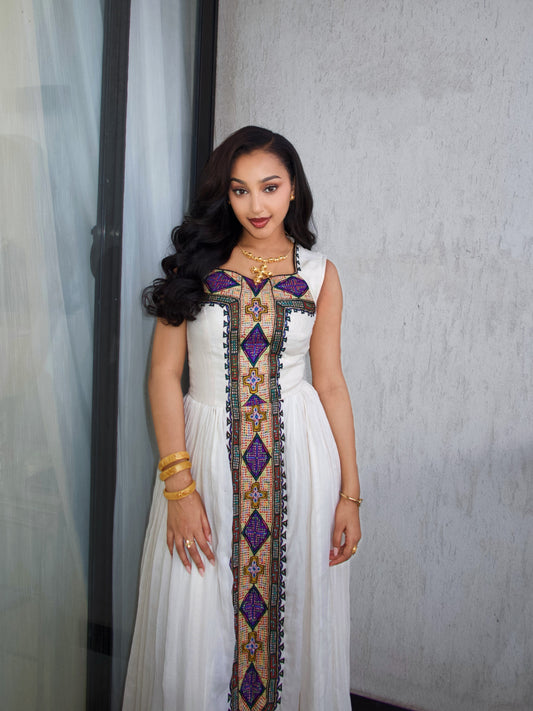 A woman wearing a sleeveless white Ethiopia Clothes with a vertical purple, gold, and green embroidered panel down the front, standing indoors against a textured light gray wall.