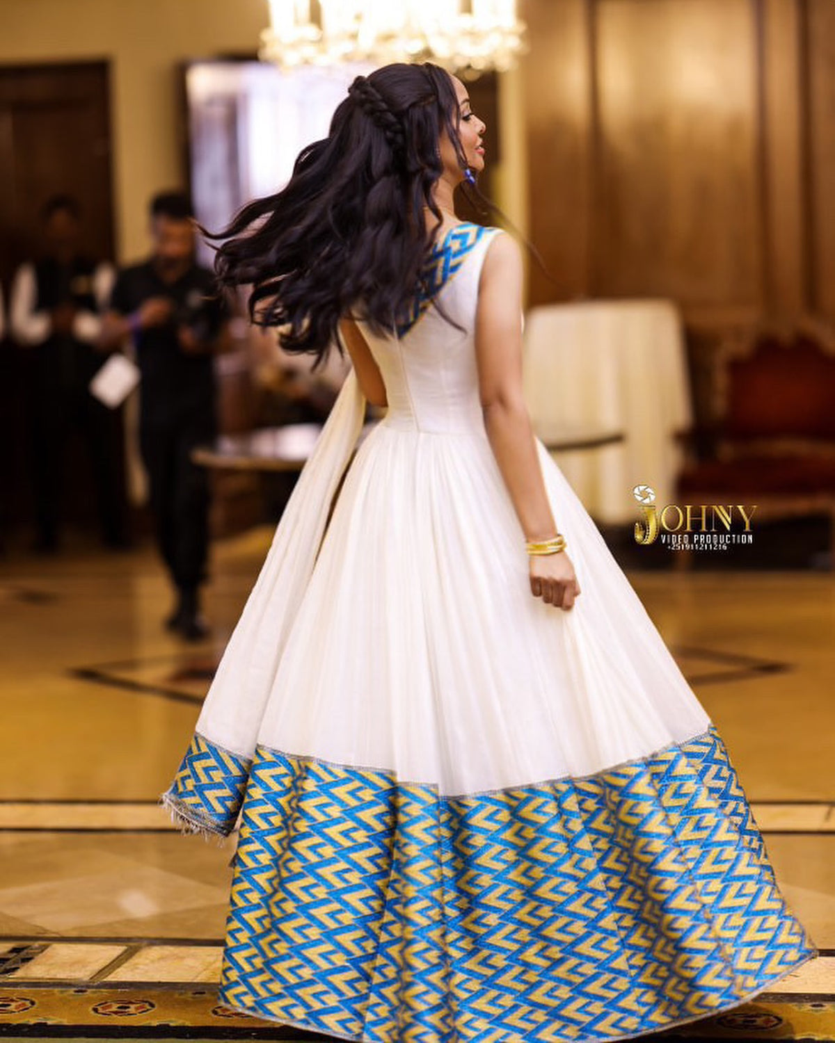 A woman wearing a white sleeveless Habesha Kemis  with a voluminous skirt featuring a blue and gold geometric pattern at the hem, photographed in an indoor event space. The brand name 'JOHNY GILLIESTOWN' is visible as a watermark on the right side.