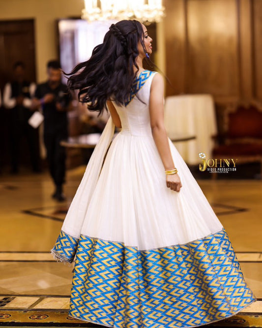 A woman wearing a white sleeveless Habesha Kemis  with a voluminous skirt featuring a blue and gold geometric pattern at the hem, photographed in an indoor event space. The brand name 'JOHNY GILLIESTOWN' is visible as a watermark on the right side.