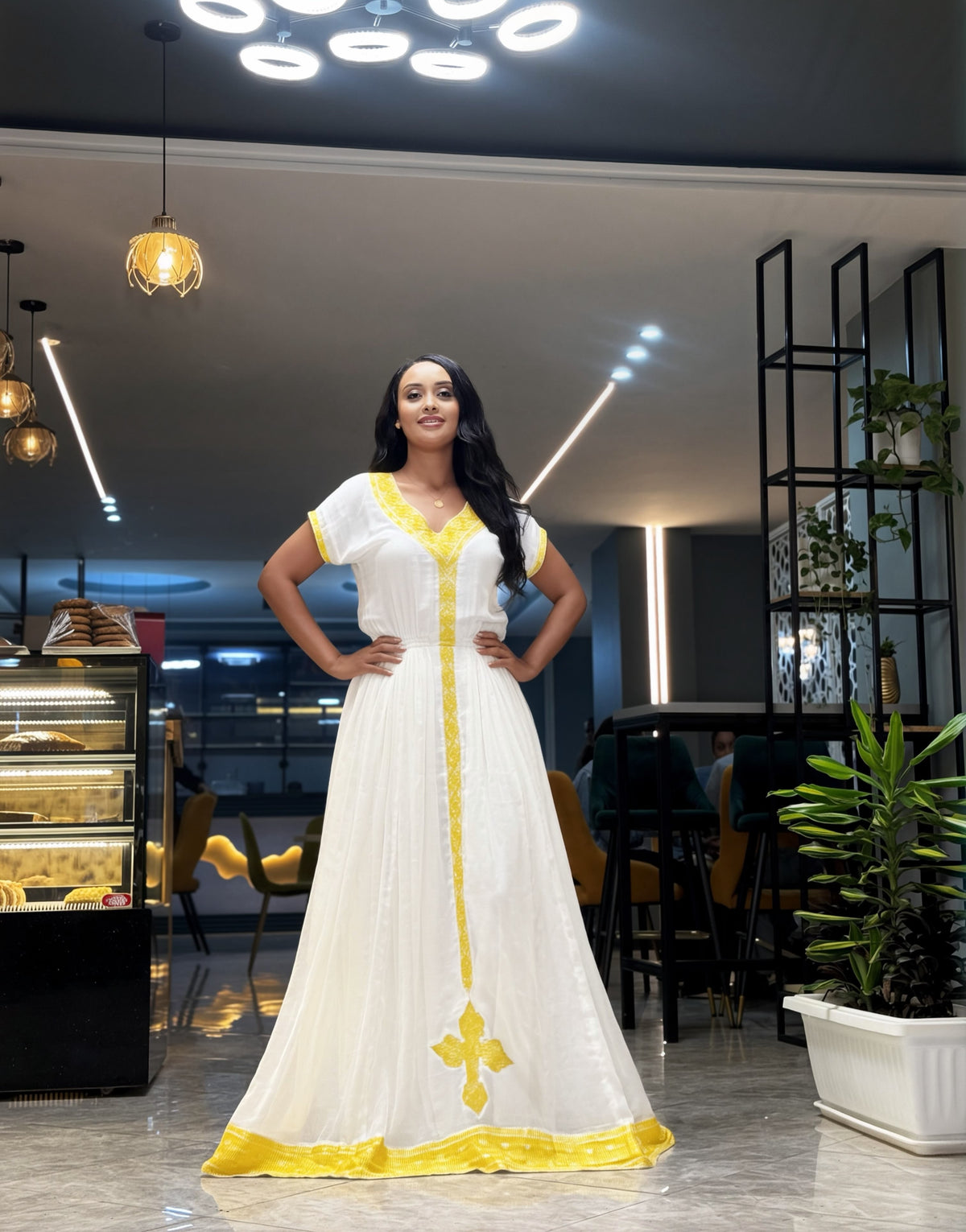 A woman stands in a modern indoor setting wearing a long white Habesha Kemis with bright yellow trim and a yellow cross motif near the hem. The background shows a contemporary interior with hanging lights, shelving, and plants.