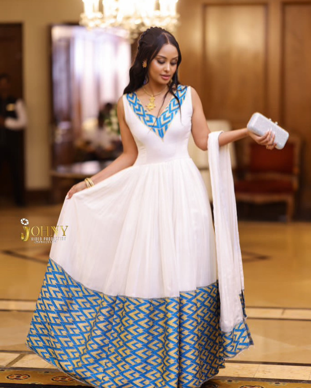 A woman wearing a white sleeveless Anarkali-style Habesha Kemis  with a blue geometric patterned neckline and a matching blue patterned flared hem, holding a white dupatta in an indoor event space.