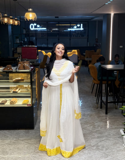 A woman wearing a white traditional Habesha Kemis with gold trim stands in a modern cafe setting with tiled floors, pastry displays, and seating in the background.