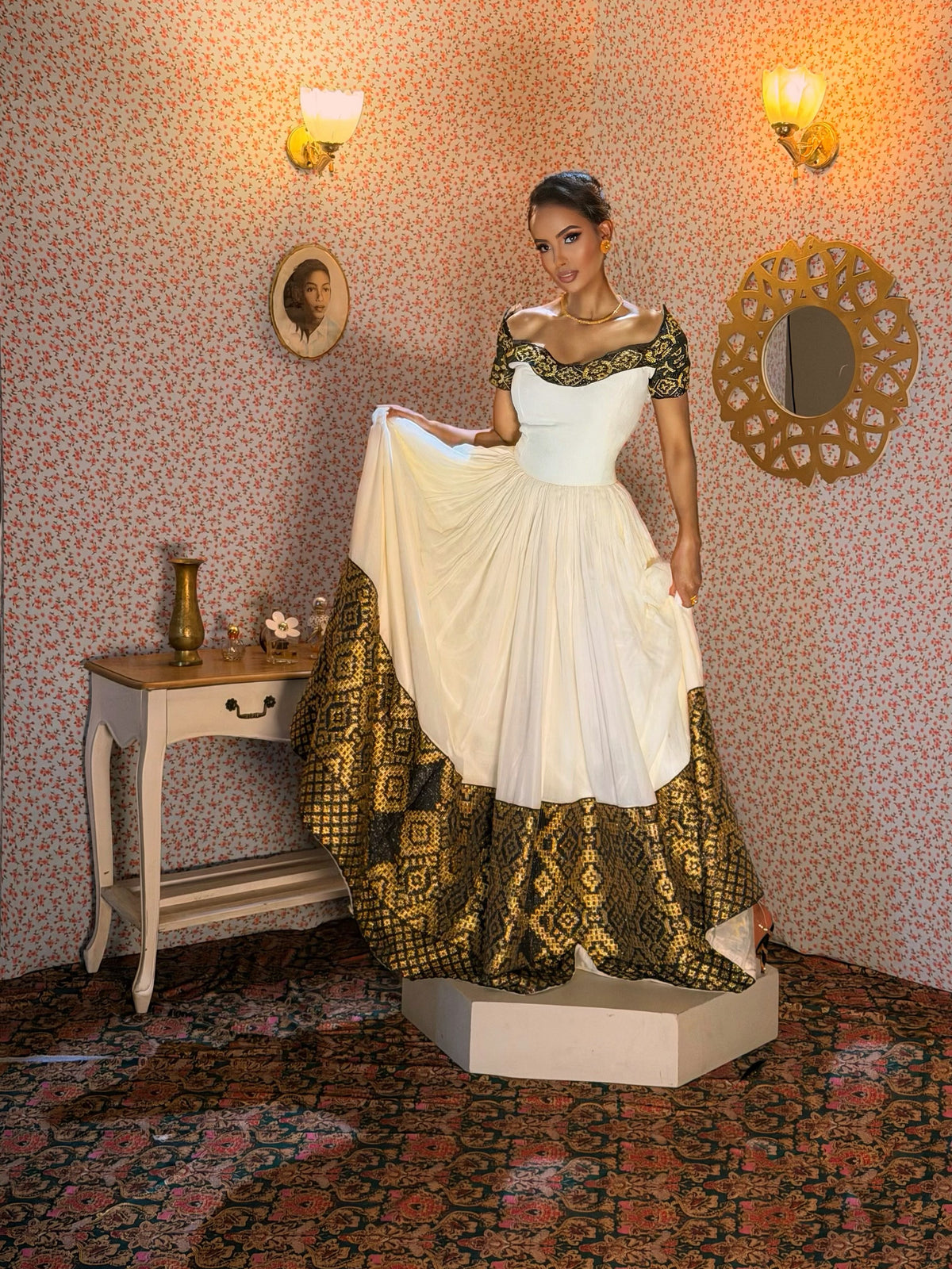A woman models a traditional Indian Ethiopian Dress with a white pleated skirt and a black and gold patterned border, standing on a pedestal in a room with patterned wallpaper and mirrors.