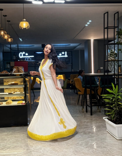 A woman wearing a flowing white Habesha Kemis with yellow trim stands in a modern cafe interior. The dress has a deep V-neck, sleeveless design, and a flared skirt with yellow border detailing.