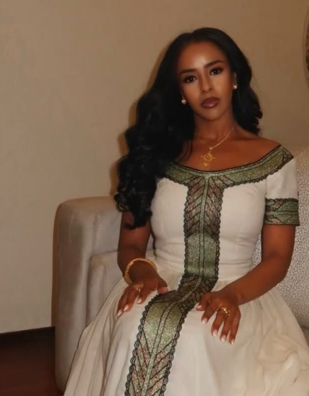 A woman with long, wavy black hair is seated indoors wearing a white Ethiopia store with green and gold embroidered details. She is wearing gold jewelry, including a necklace, earrings, and bracelets, and is sitting on a light-colored upholstered armchair against a plain beige wall.