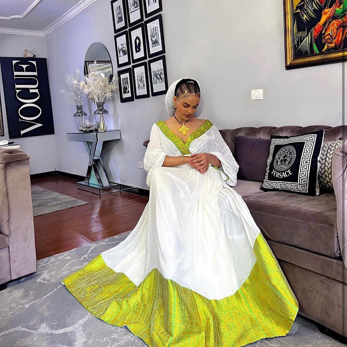 A woman wearing a traditional white Habesha Dress with a bright yellow-green border and matching head covering, seated on a brown sofa in a living room. A Versace pillow is visible on the sofa, and a Versace wall sign appears in the background.