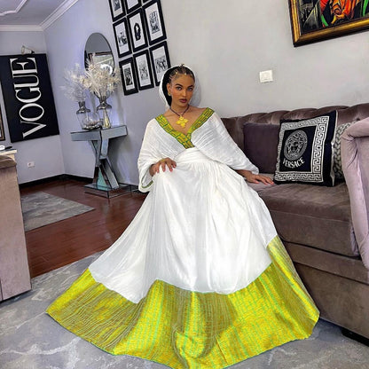 A woman wearing a voluminous white traditional Habesha Dress with a bright lime green V-neckline and a matching lime green patterned hem sits on a brown sofa in a living room. She is wearing gold jewelry, including a prominent necklace.