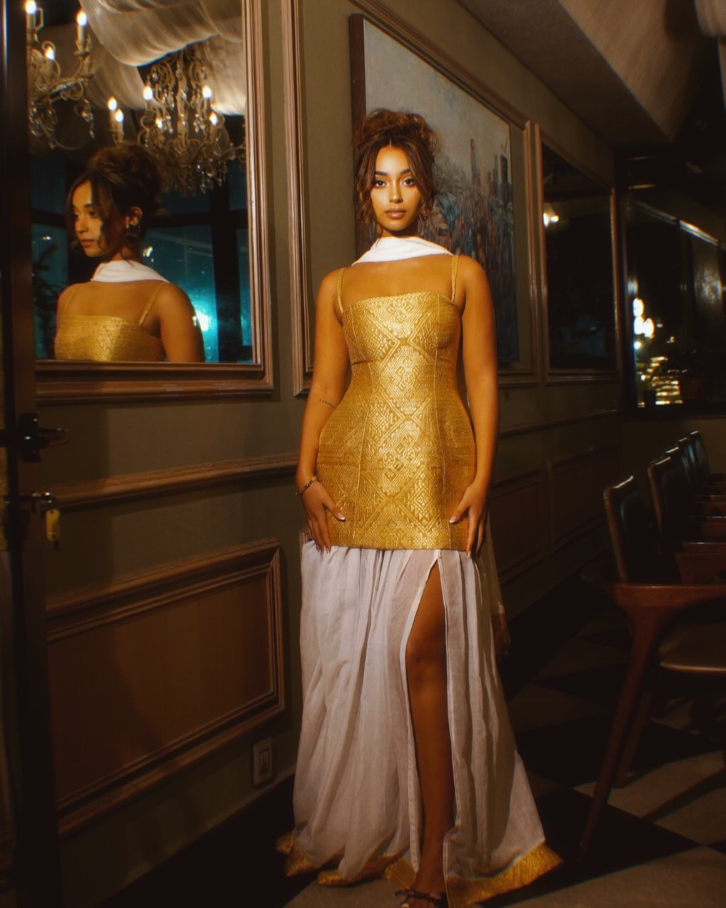 A woman stands indoors wearing a sleeveless fitted gold Habesha Dress  with a structured bodice and a flowing white skirt featuring a high slit. The setting appears to be a dimly lit interior with framed wall art, mirrors, and warm ambient lighting.