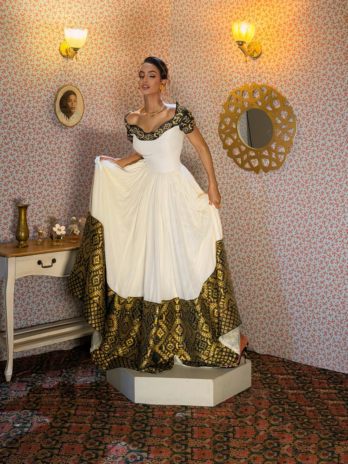 A woman models a white off-the-shoulder ball Ethiopian Dress with black lace detailing on the bodice and sleeves, and a dramatic floor-length skirt featuring a wide black and gold patterned border. She stands on a white pedestal in a vintage room with patterned wallpaper, a side table, and a patterned rug.