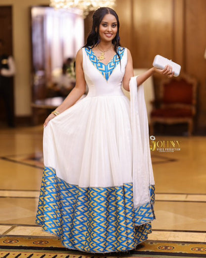 A woman modeling a white sleeveless Habesha Kemis with a blue and gold geometric patterned neckline and flared hem, paired with a matching white dupatta in an indoor event setting.