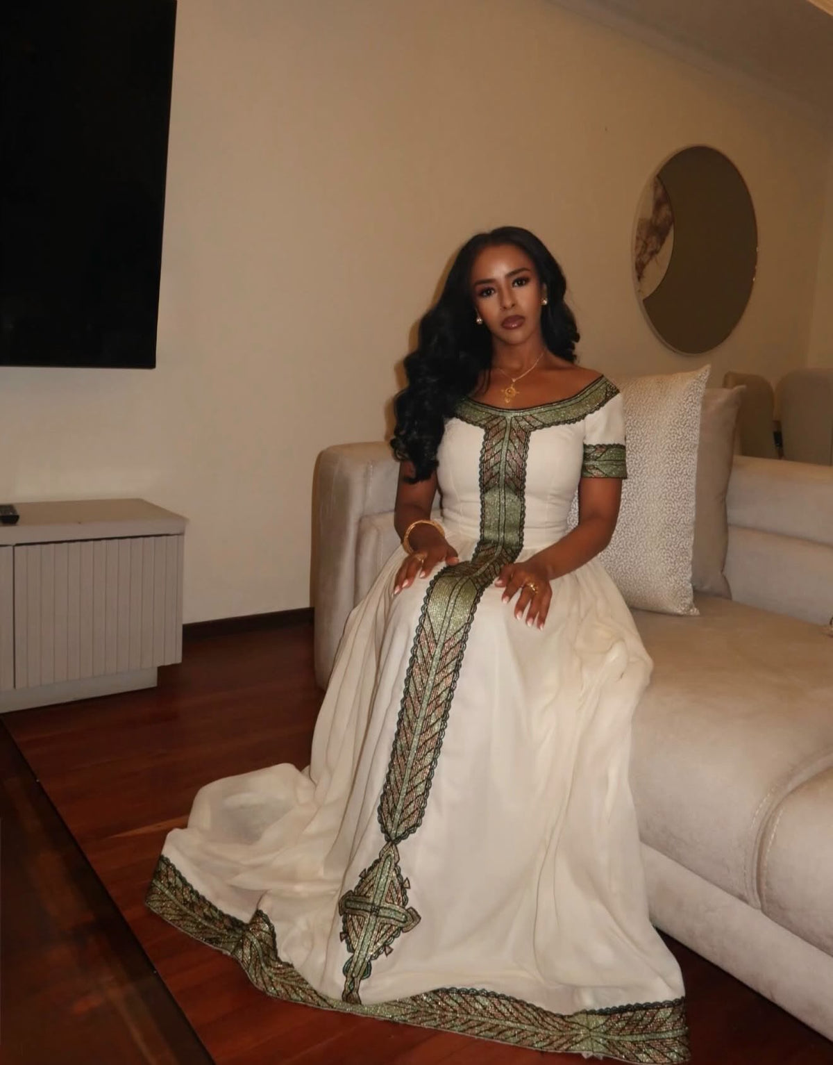 A woman wearing a long white Ethiopia store with green and gold patterned trim sits on a beige sofa in a living room.