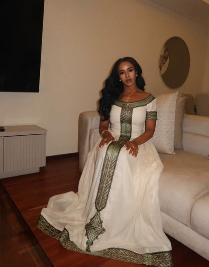 A woman wearing a long white Ethiopia store with green and gold patterned trim sits on a beige sofa in a living room.