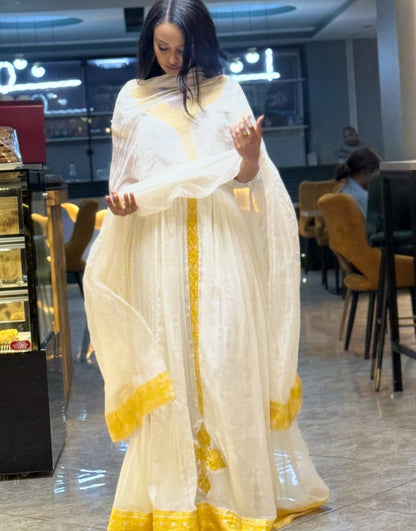 A woman wearing a white traditional Habesha Kemis with yellow embroidered borders, standing in a restaurant or cafe interior.
