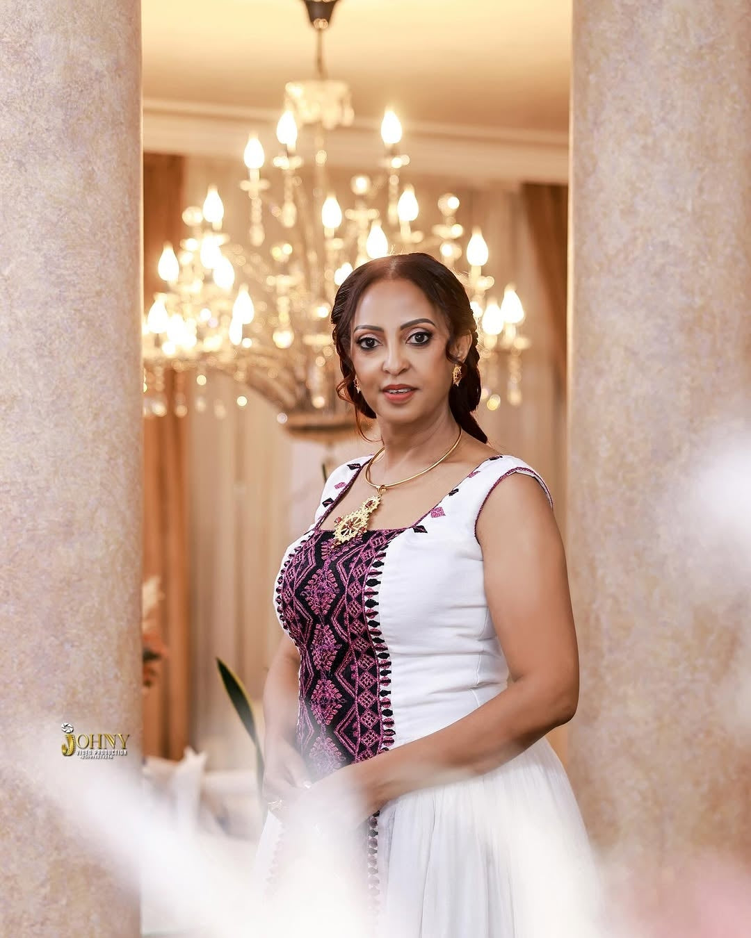 Woman in a white and Purple Habesha Kemis standing in a decorated room with chandeliers.