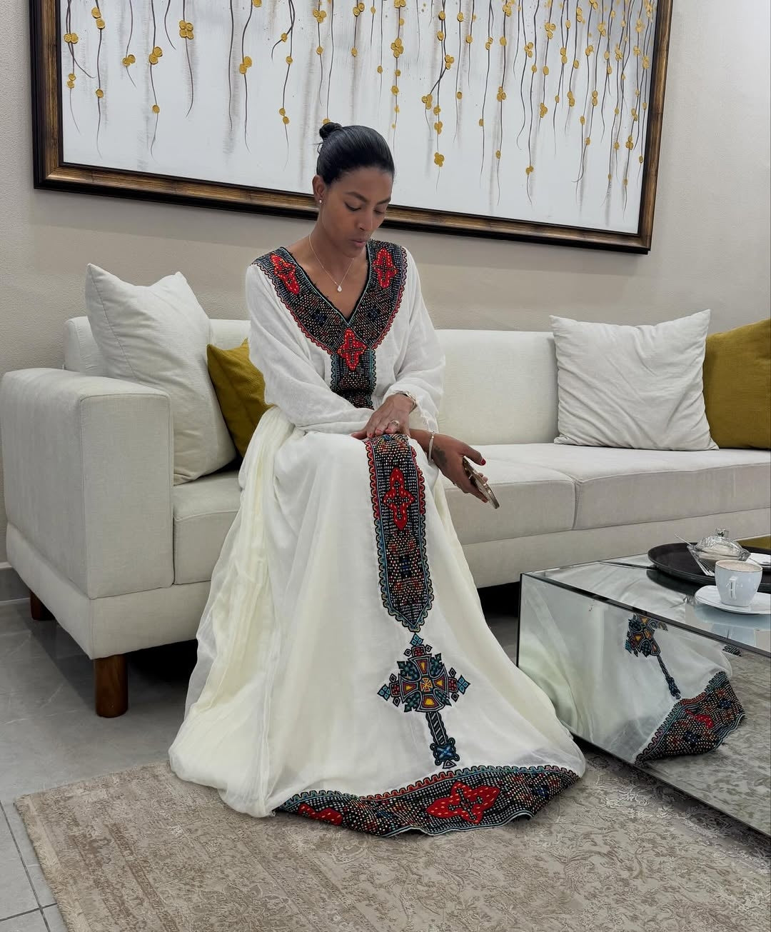 Woman in a white Habesha dress  with red and blue patterns sitting on a couch in a living room.
