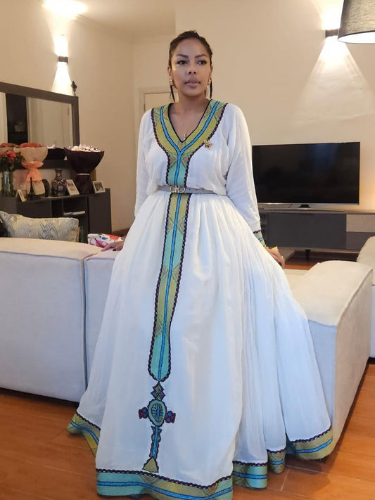 Woman in a Habesha Zuria with blue and green accents standing in a living room.