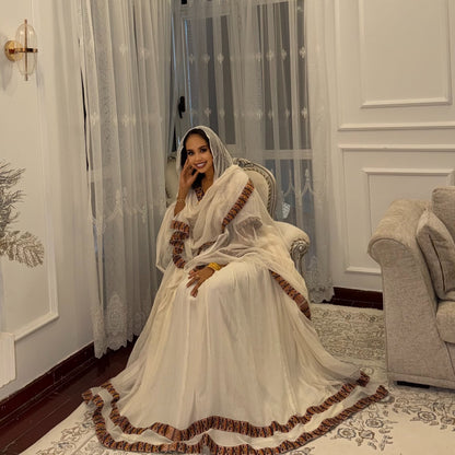 A woman wearing a white traditional Habesha Kemiswith brown ruffled trim sits in an ornate chair in a decorated room with sheer curtains, a sofa, and a patterned rug.