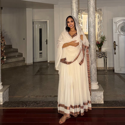 A woman wearing a traditional white or cream-colored Habesha Kemiswith brown patterned borders and a matching head covering, standing in an elegant indoor hallway with columns and tiled floors.