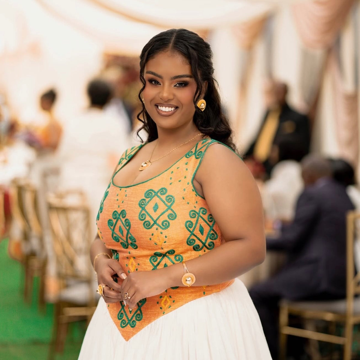 A woman smiling in a vibrant orange and green patterned sleeveless top with a white Habesha Dress , standing in a softly blurred event setting.