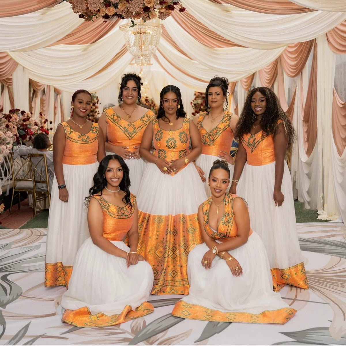 Seven women posing together in matching formal Habesha Dress  with orange and gold embroidered bodices and white flared skirts, set against a decorated event backdrop with draped cream fabric and a chandelier.