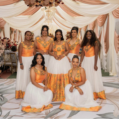 Seven women posing together in matching formal Habesha Dress  with orange and gold embroidered bodices and white flared skirts, set against a decorated event backdrop with draped cream fabric and a chandelier.
