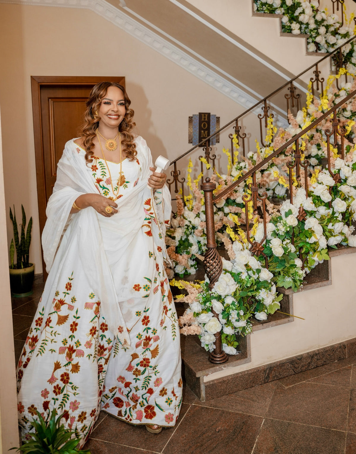 A woman wearing a white traditional Ethiopian Traditional Dress with colorful floral embroidery from Tsehaye Mehari stands on an indoor staircase decorated with white and yellow flowers. She is holding a small white item in her hand.