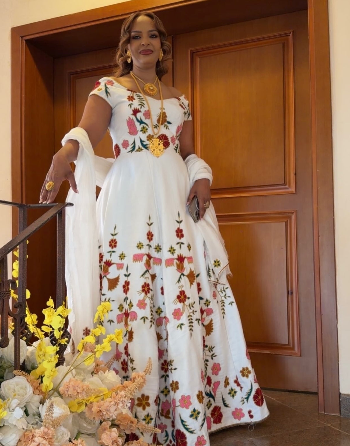 A woman wearing a white off-the-shoulder Ethiopian Traditional Dress with colorful floral embroidery, standing in front of a wooden door.