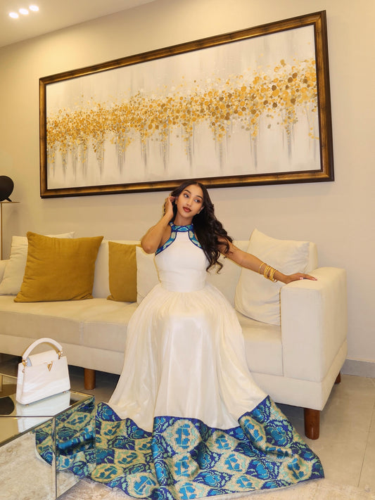 Woman in a white dress with blue Ethiopian Traditional Dresssitting on a couch in a living room.