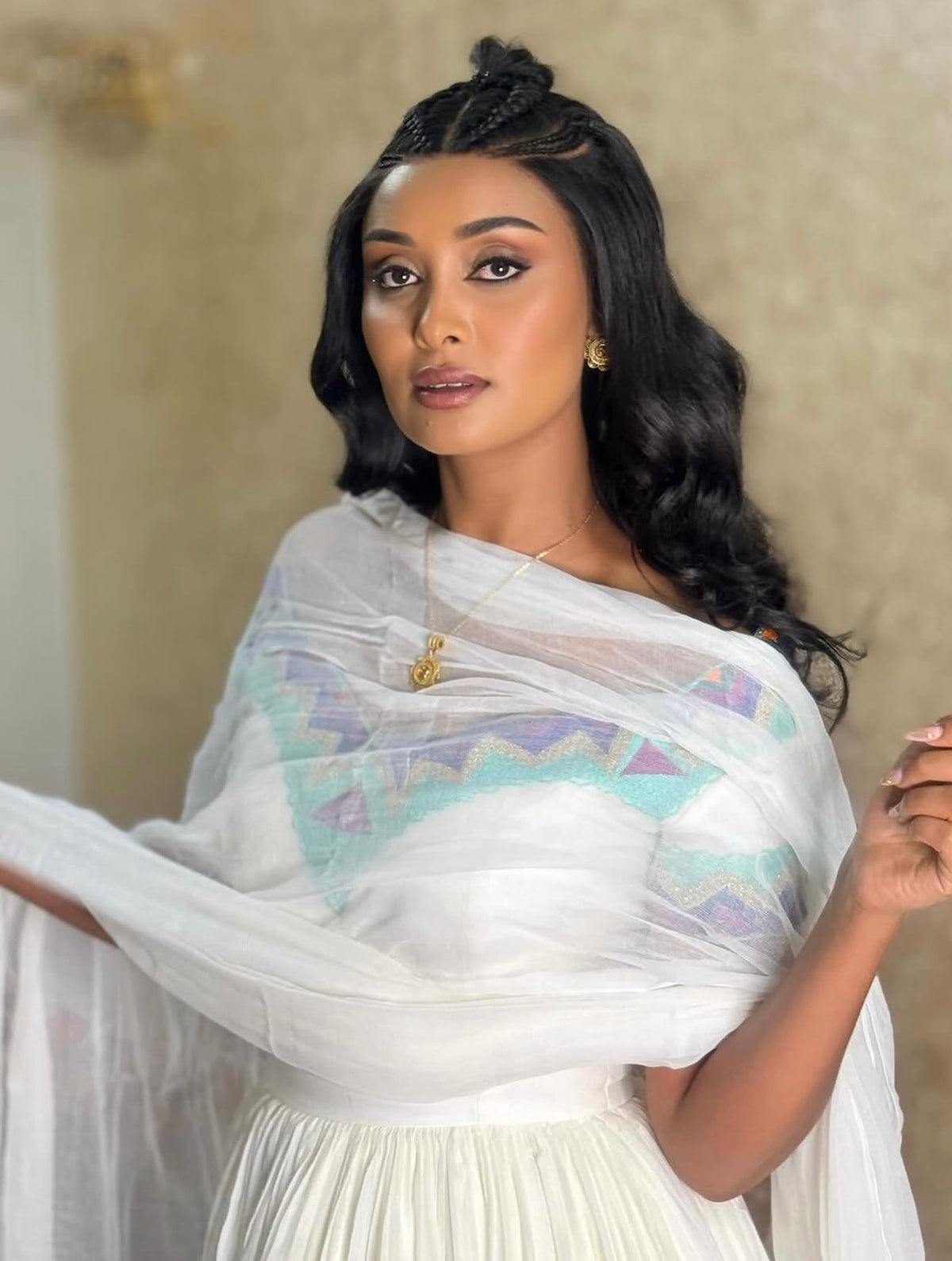 A woman wearing a white traditional  Ethiopian Dress with a sheer white shawl featuring a pastel geometric pattern in light blue, purple, and green, posed against a neutral beige background.