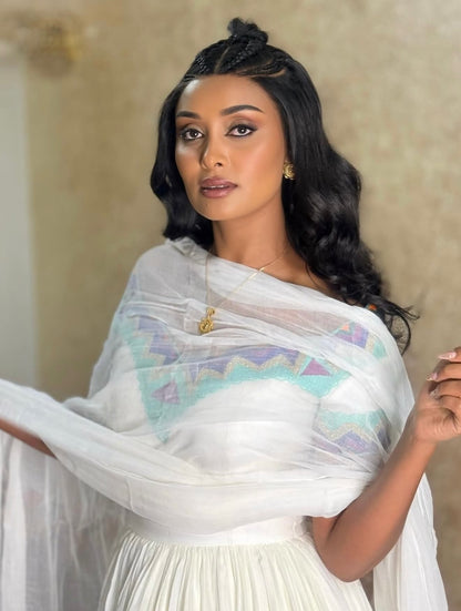 A woman wearing a white traditional  Ethiopian Dress with a sheer white shawl featuring a pastel geometric pattern in light blue, purple, and green, posed against a neutral beige background.