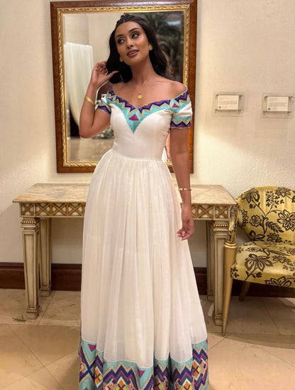 A woman modeling a white off-the-shoulder  Ethiopian Dress  with colorful geometric patterned trim at the neckline and hem, standing in an indoor room with a mirror, console table, and patterned chair.