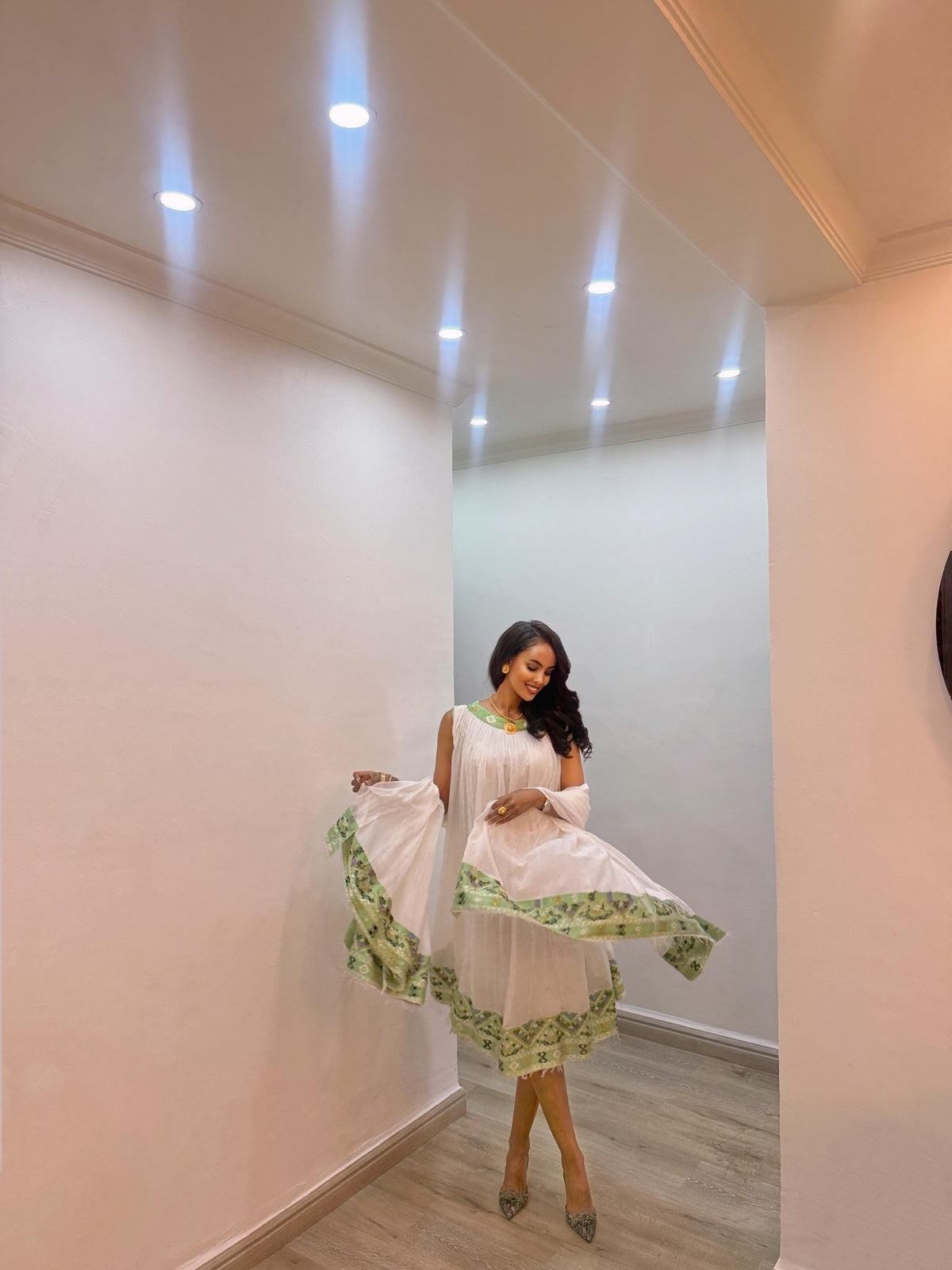 A woman modeling a white sleeveless Habesha Kemis with green patterned ruffled trim, standing in a brightly lit indoor hallway with recessed ceiling lights.