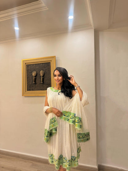 A woman wearing a white sleeveless Habesha Kemiswith green printed borders and a matching green dupatta, standing in an indoor room with cream walls and a framed picture.
