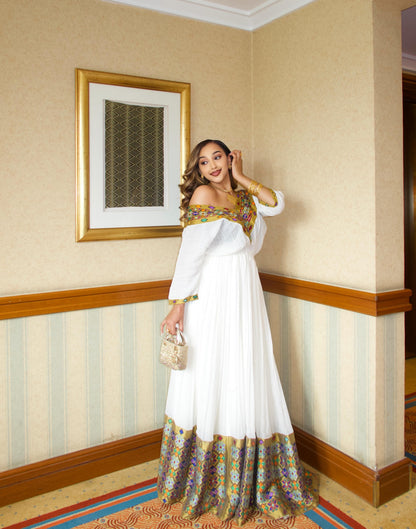 A woman models a white off-the-shoulder maxi Traditional Ethiopian dress with gold and green patterned embroidery, standing in a hotel hallway with beige walls and striped wallpaper.