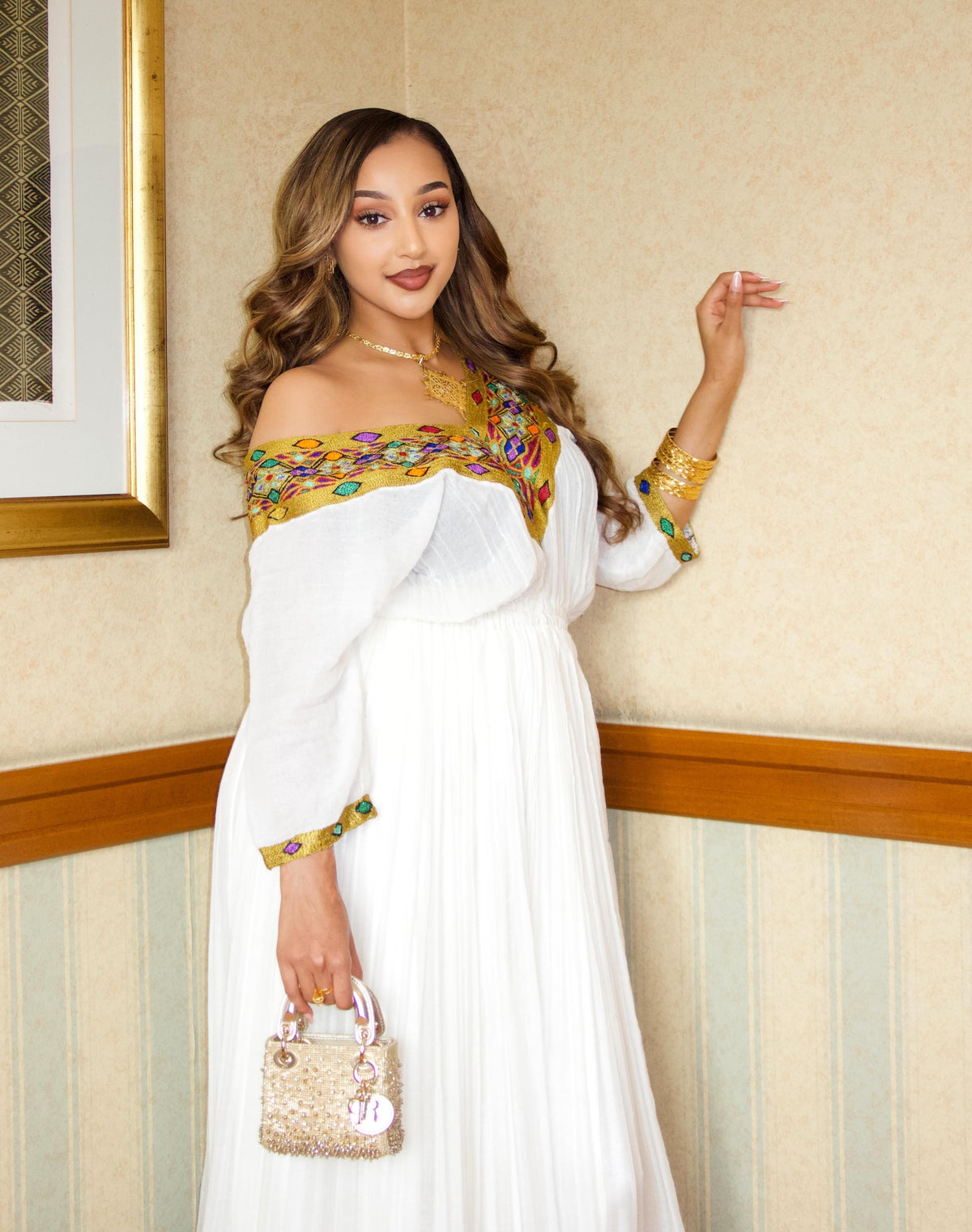 A woman models a white off-the-shoulder maxi Traditional Ethiopian dress with gold and multicolor embroidered detailing on the bodice and cuffs, holding a small gold handbag in an indoor setting.