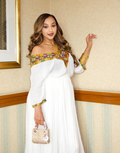 A woman models a white off-the-shoulder maxi Traditional Ethiopian dress with gold and multicolor embroidered detailing on the bodice and cuffs, holding a small gold handbag in an indoor setting.