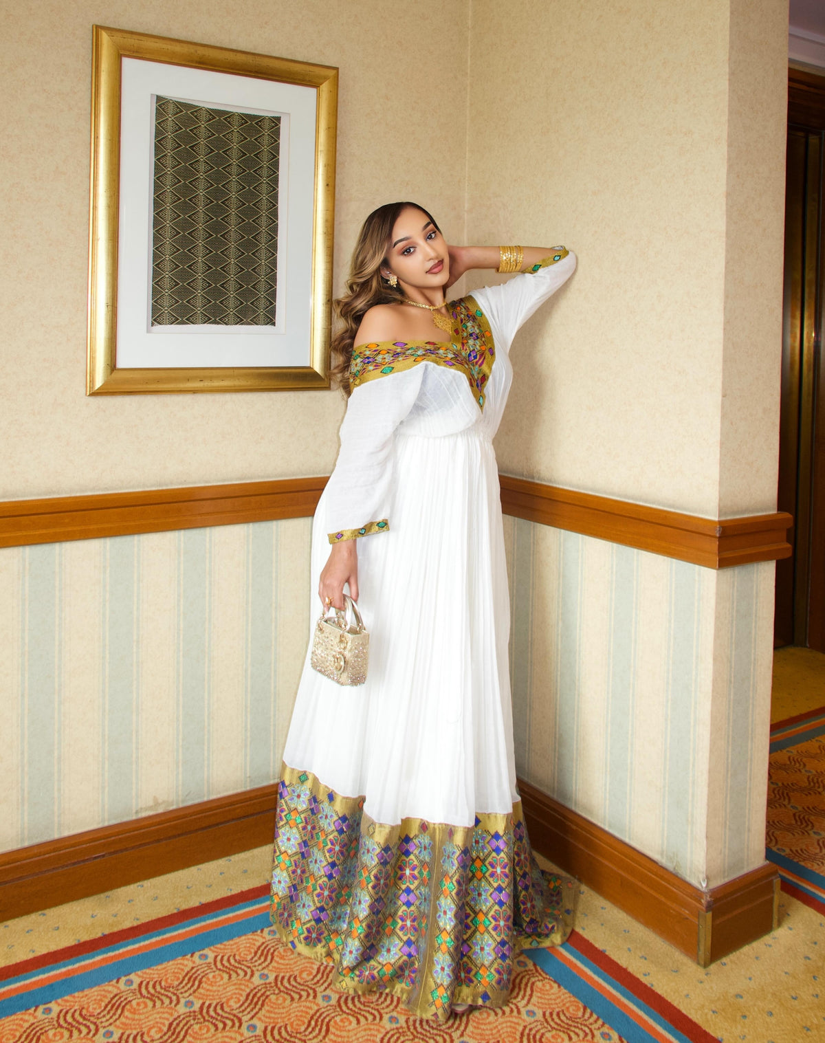 A woman models a white off-the-shoulder maxi Traditional Ethiopian dress with gold, green, and purple patterned borders, standing in a hotel hallway next to a beige wall with wood trim.