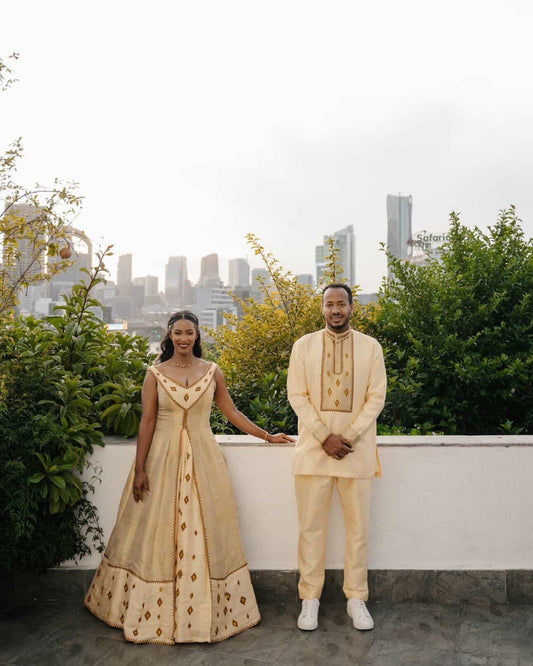 A couple posing in matching traditional Indian Habesha matching couple outfit on a rooftop terrace with a city skyline and greenery in the background. The woman wears a cream/beige sleeveless gown with gold embroidery, and the man wears a matching cream/beige kurta and trousers set.