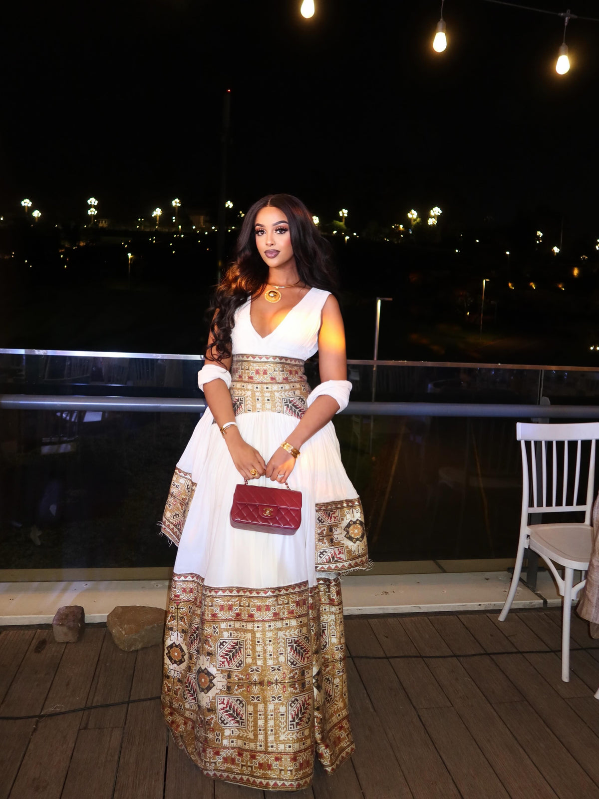 Woman in a white and patterned Habesha Dress holding a red clutch on a wooden deck at night.